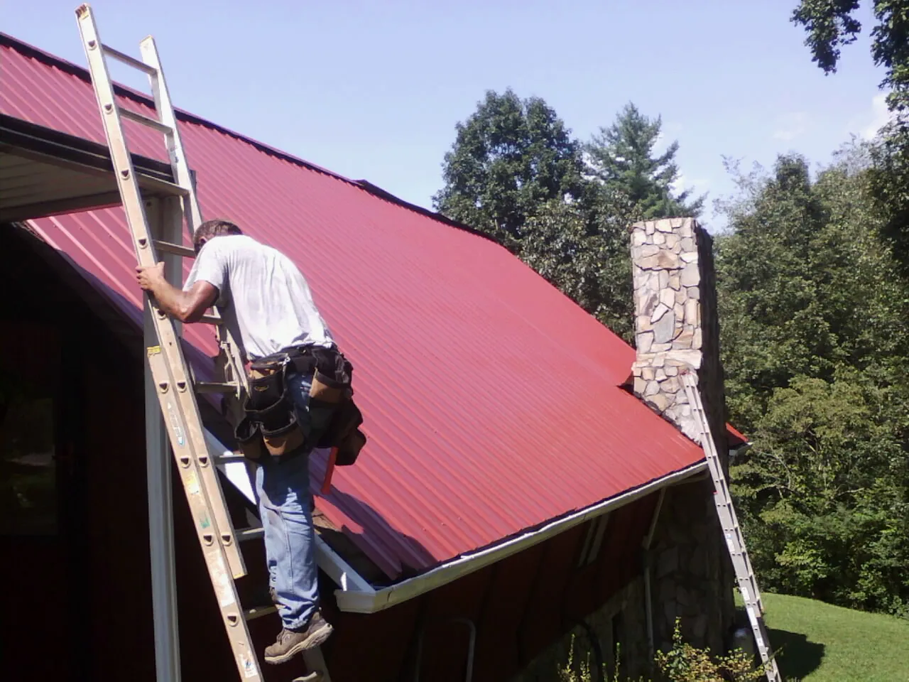 Roofer on a ladder beside a red metal roof and stone chimney — Soffit & Fascia Repair by Roofing Brothers in Monclova